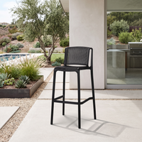 A black outdoor bar stool with a woven seat and backrest, positioned on a light concrete patio in front of a modern house with a pool and landscaped garden in the background.