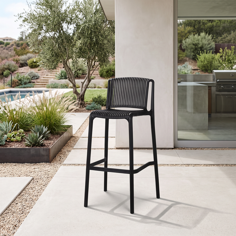 A black outdoor bar stool with a woven seat and backrest, positioned on a light concrete patio in front of a modern house with a pool and landscaped garden in the background.