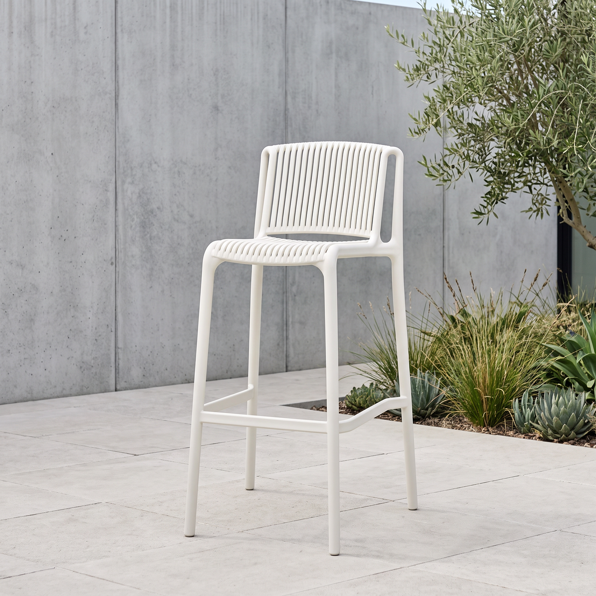 A modern white outdoor bar stool with a slatted plastic seat and backrest, positioned on a light stone patio against a gray concrete wall with green plants.